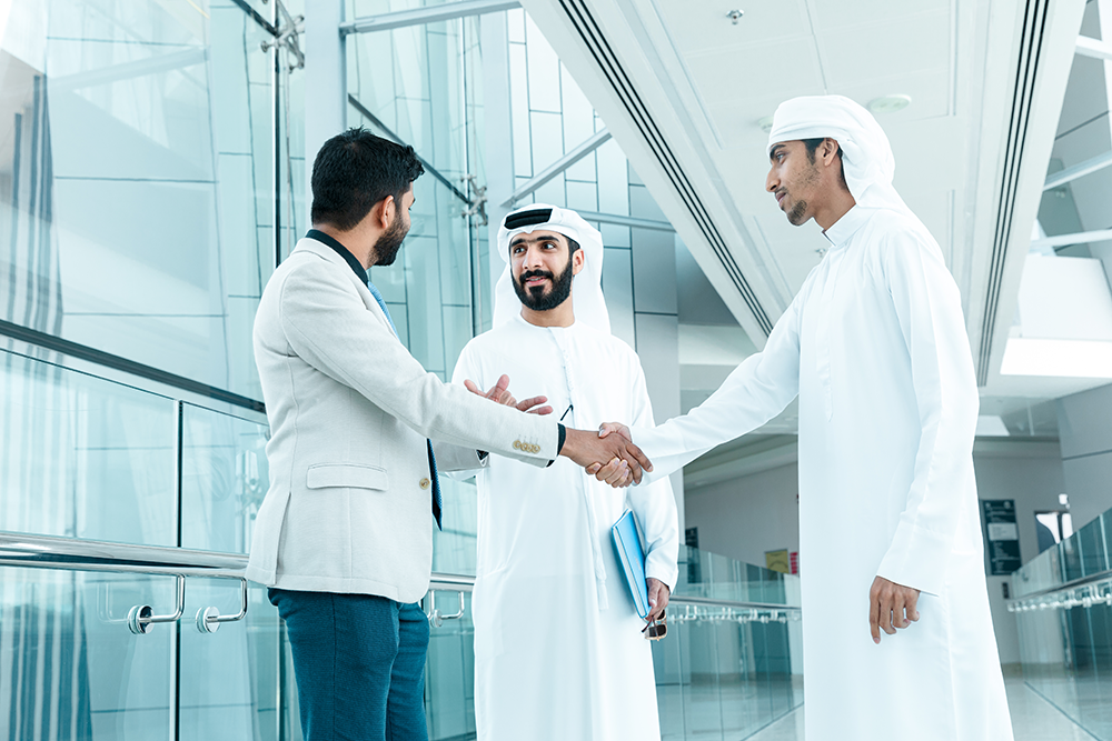 Three professional men shaking hands and conversing in a modern glass corridor.