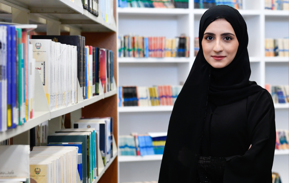 A female student posing in a library with rows of books.