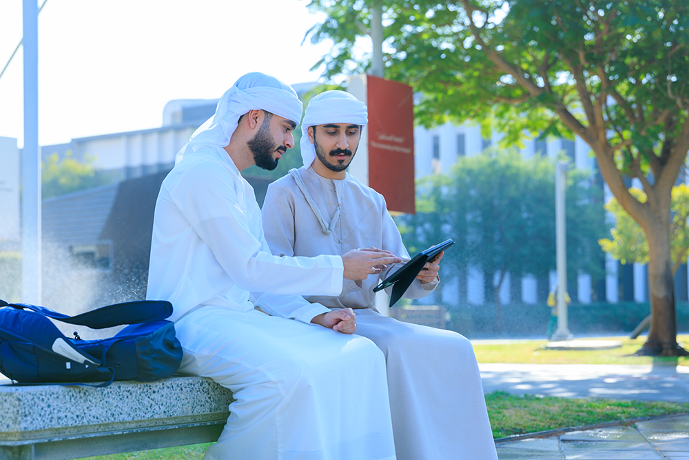 Two male students in traditional attire sitting on a campus bench and reviewing content on a tablet.