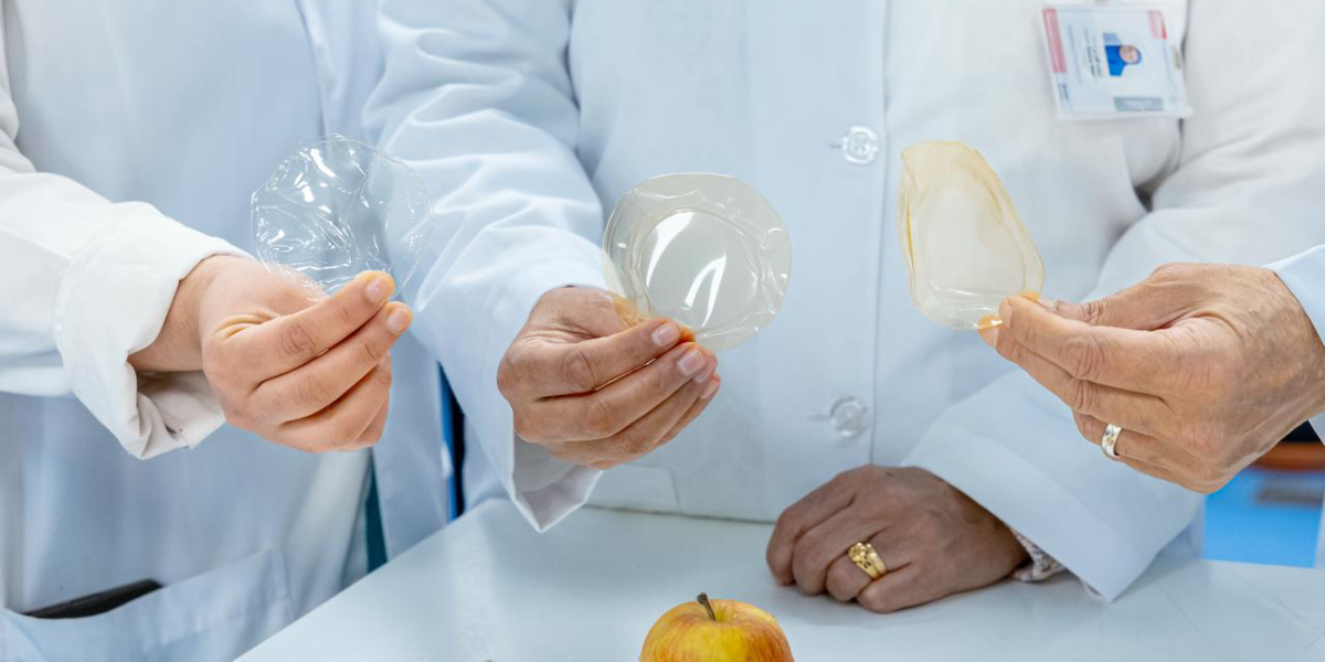 Three people in lab coats hold different samples of a thin, transparent edible food-preservation film in their hands, demonstrating the innovative product designed to maintain the freshness of produce. An apple rests on the table in front of them, symbolizing the type of fresh food the film helps preserve. This image relates to UAEU securing a U.S. patent for this sustainable edible film that reduces food waste.