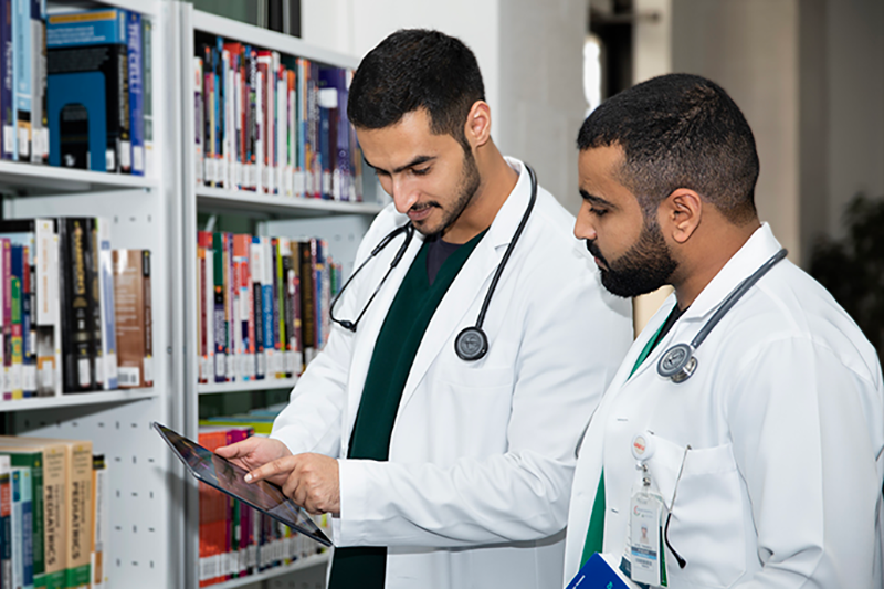 Two male medical students in white lab coats and stethoscopes reviewing information on a tablet in a library setting.