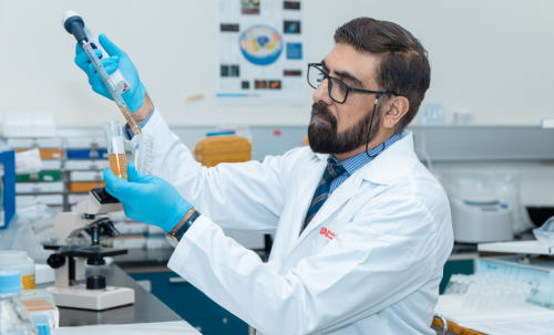 عالم يرتدي معطف مختبر وقفازات ونظارات واقية يستخدم ماصة لنقل سائل إلى أنبوب اختبار داخل مختبر في المركز الوطني للمياه والطاقة بجامعة الإمارات العربية المتحدة