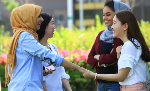 أربع طالبات متنوعات يتحدثن ويتصافحن في حرم جامعي أخضر، مما يعكس الصداقة والشمول والرفاهية بما يتماشى مع مركز الإمارات لأبحاث السعادة.