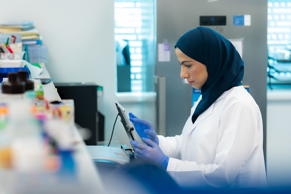 باحثة ترتدي معطف المختبر وقفازات زرقاء تستخدم جهازاً لوحياً لمراقبة المعدات في مختبر طبي حديث.